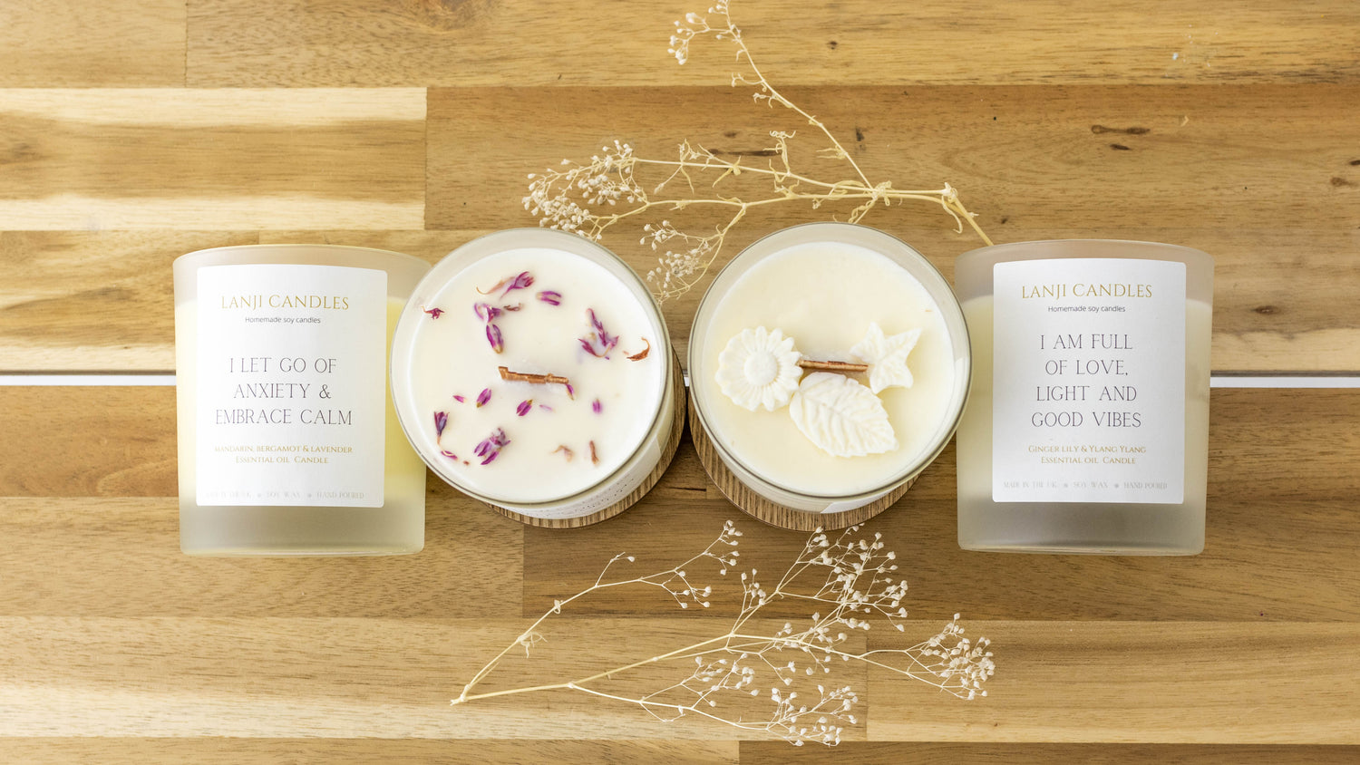 Four candles arranged on a light wood surface: two frosted glass jars with white labels and two round white candles, one with dried flower petals and one with white flower decorations. Decorative dried stems are scattered around them.